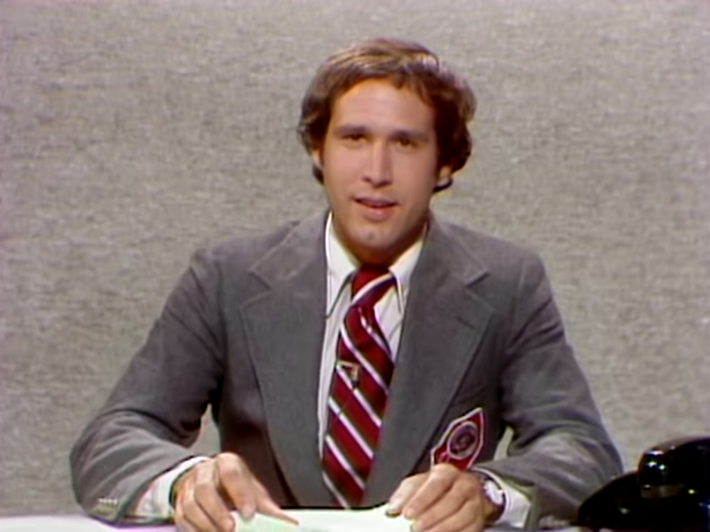 Chevy Chase performing on Saturday Night Live in a grey suit with an oversized red and white striped tie