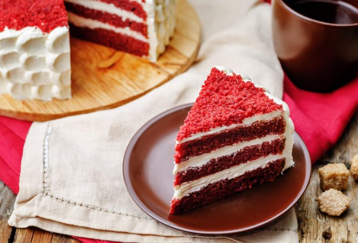 A slice of red velvet cake on a small plate with a mug of coffee and the rest of the cake in the background.
