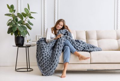 A woman sitting on a cream couch with a chunky gray knitted blanket.