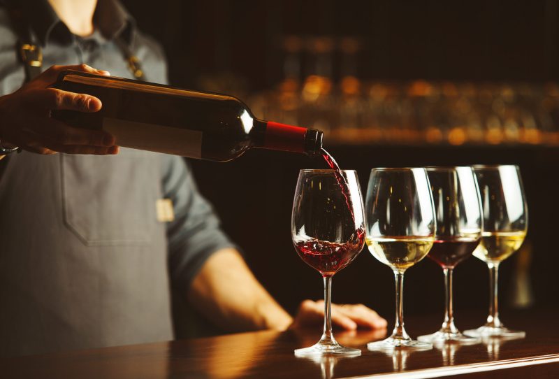 Bartender pouring wine into a glass, various varieties of wine on a wooden table.