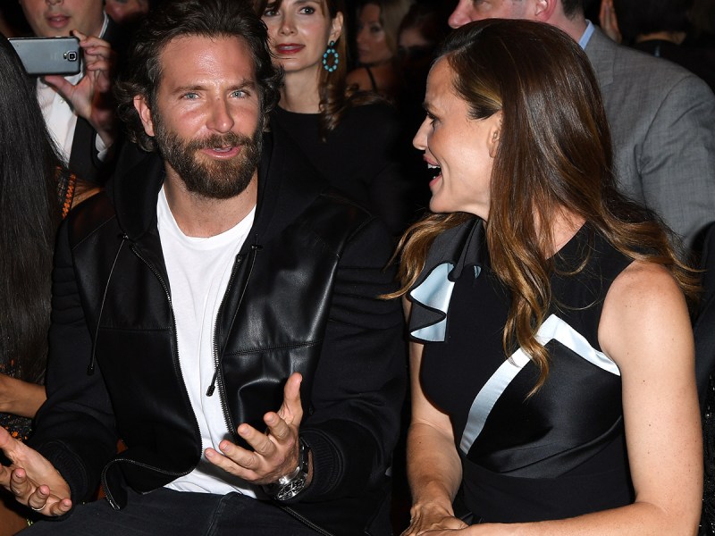 Bradley Cooper in a white t-shirt and leather jacket looking confused, sitting next to Jennifer Garner, who is facing him and laughing.