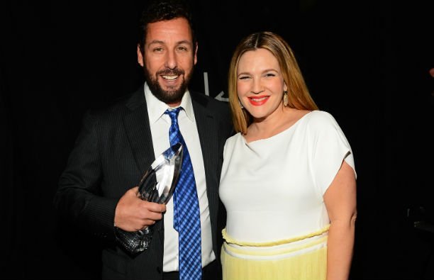 Adam Sandler wearing a dark suit standing with Drew Barrymore, who's wearing a white top and yellow