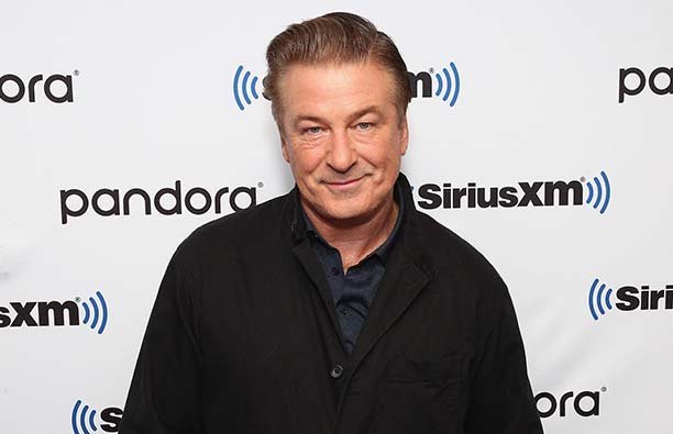 Alec Baldwin in a dark shirt and jacket smiling in front of a SiriusXM background