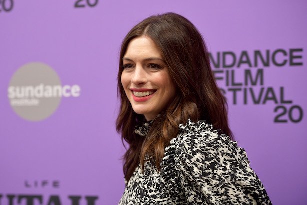 Anne Hathaway smiles in a black and white dress against a purple background at Sundance 2020