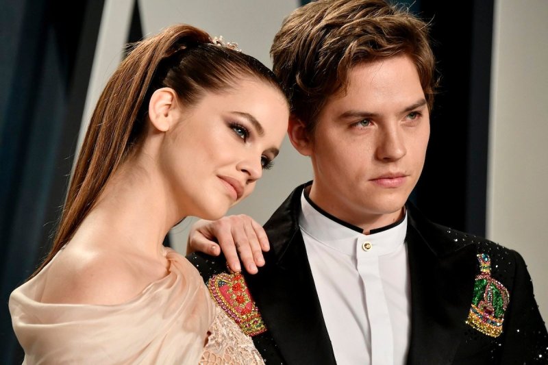 Barbara Palvin in a tan dress resting her hand on the shoulder of Dylan Sprouse in a black suit/whit