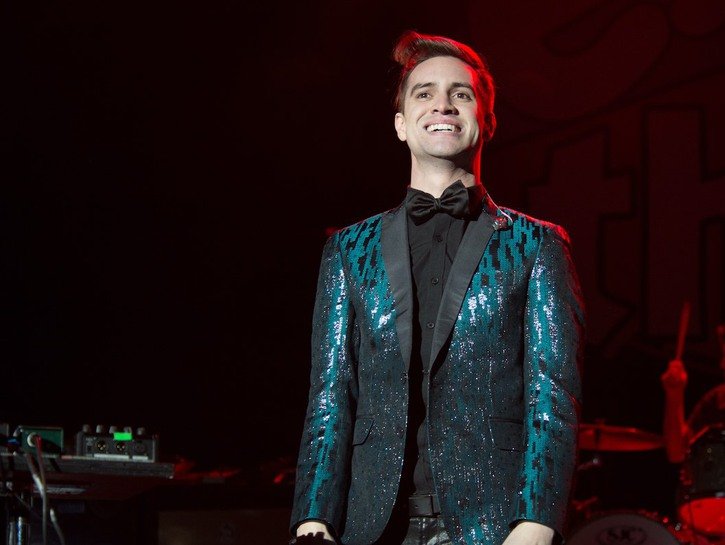 Brendon Urie of Panic! At The Disco in a blue suit smiles on a dark stage