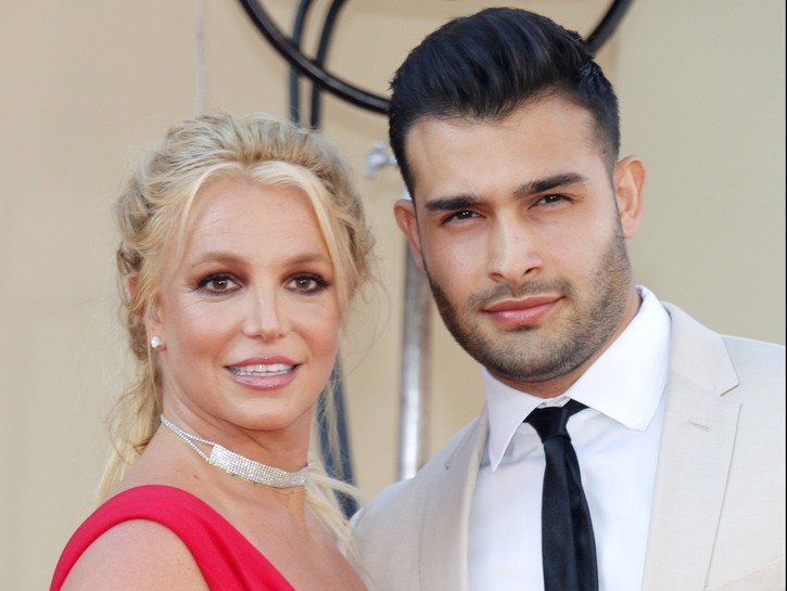 Britney Spears, in red, standing with Sam Asghari, wearing a cream colored suit, at a movie premier