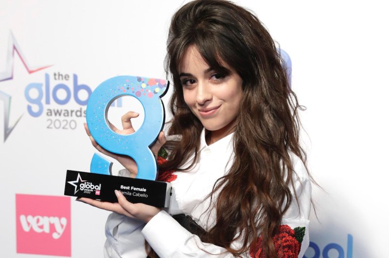 Camila Cabello in a white shirt with rose patches smiles and holds up her g-shaped award