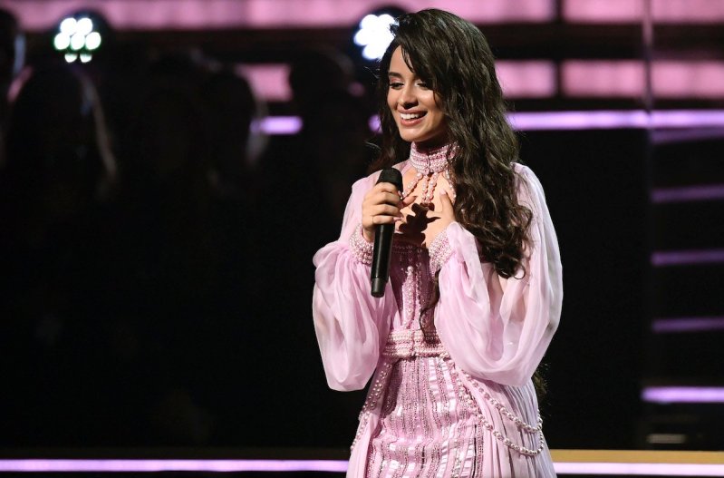Camila Cabello smiles while holding both arms to her chest while holding a mic in a pink dress