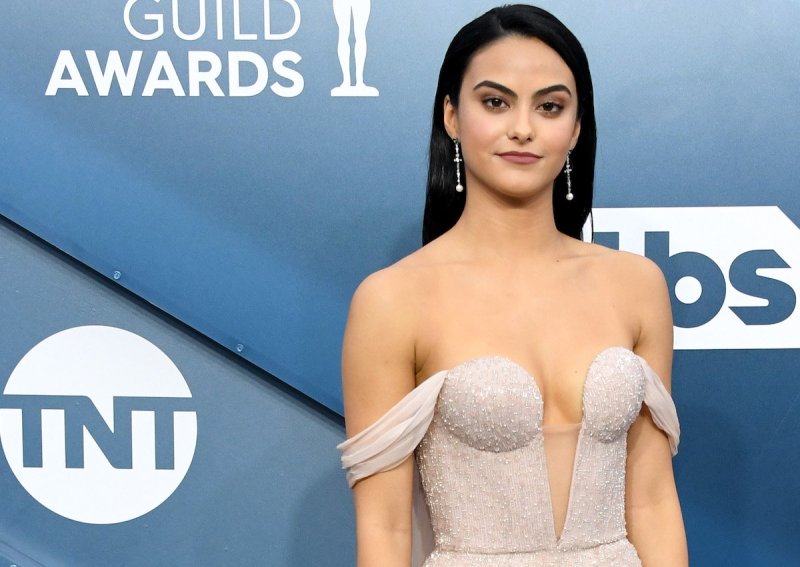 Camila Mendes smiles at the camera in champagne colored dress against a blue background