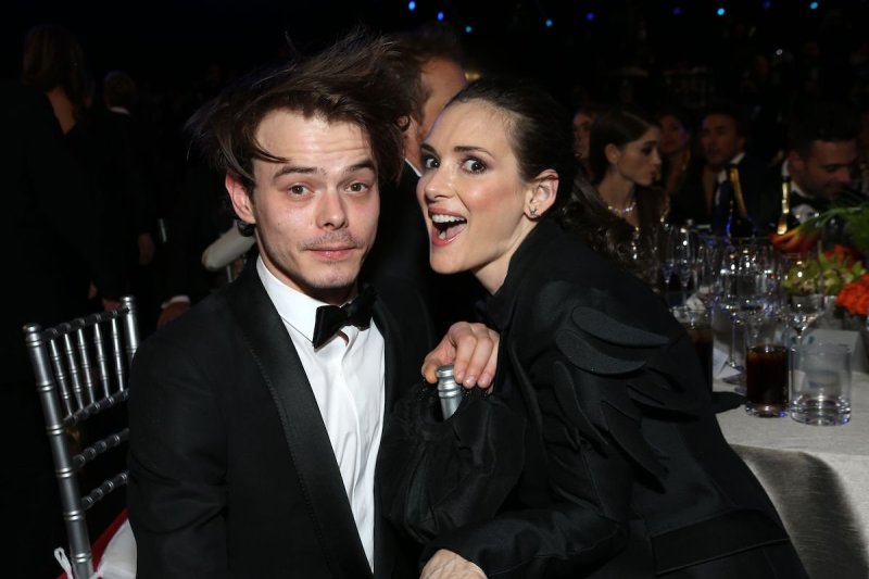 Charlie Heaton smiles in a tuxedo with Winona Ryder leaning close to him and laughing in a black dre