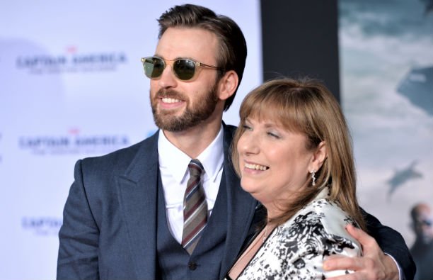 Chris Evans in a navy suit standing with his mom, Lisa, in a white floral print top on the red carpe