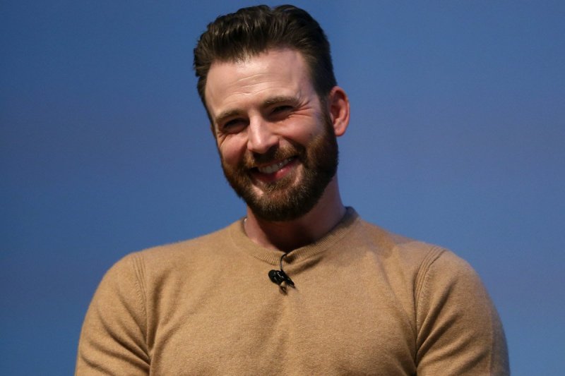 Chris Evans smiles and tilts his head dressed in a brown sweater against a blue background