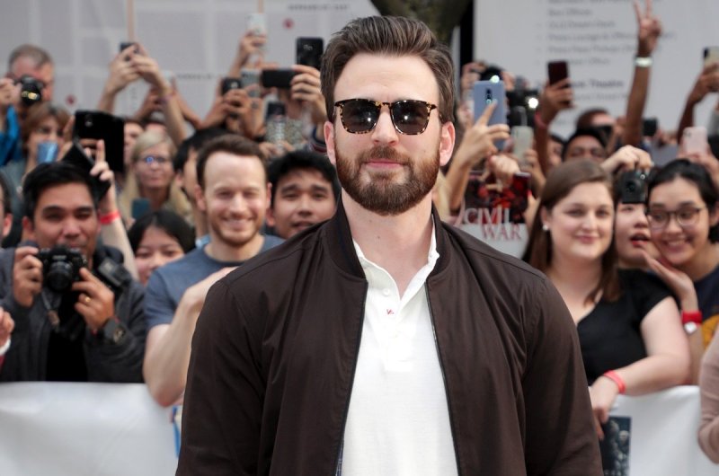 Chris Evans smiles with brown sunglasses, a brown bomber jacket, white polo, and patterned pants