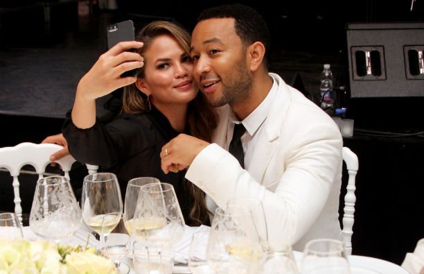 Chrissy Teigen in a black blouse and John Legend in a white suit taking a selfie at the White Party