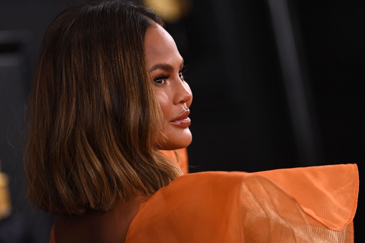 Chrissy Teigen in an orange dress looking over her shoulder before the Grammys
