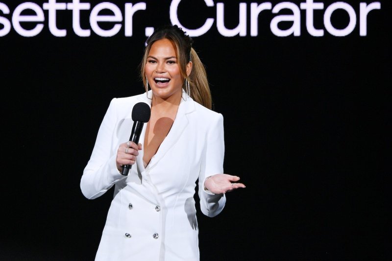 Chrissy Teigen laughs holding a mic dressed in a white pantsuit against a black background
