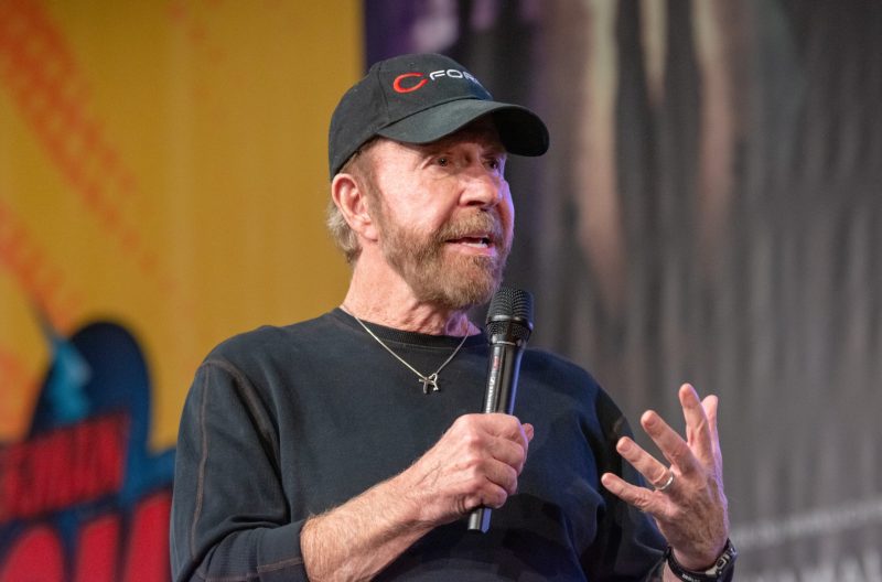 Chuck Norris wears a dark sweatshirt and holds a microphone as he addresses a crowd.