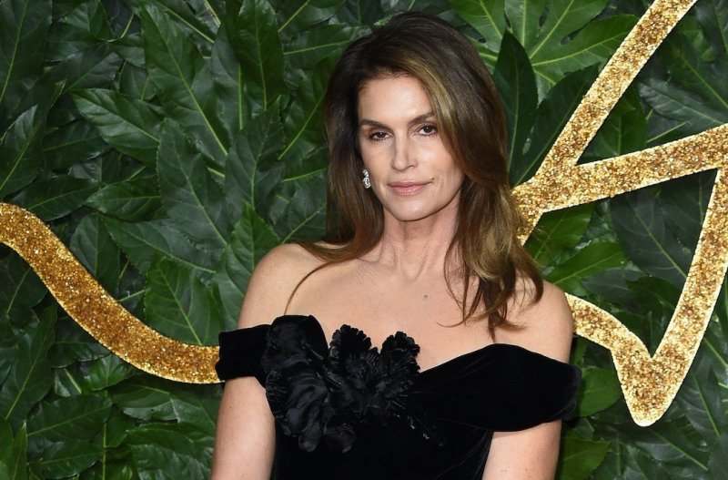 Cindy Crawford smiles at the camera in a black dress against a green and gold background