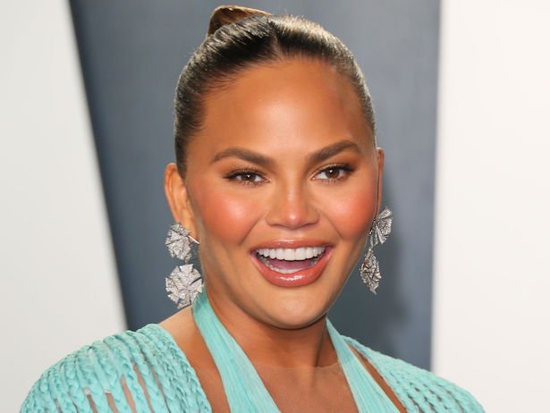 close up of Chrissy Teigen smiling and laughing in a light blue dress on a white and grey background