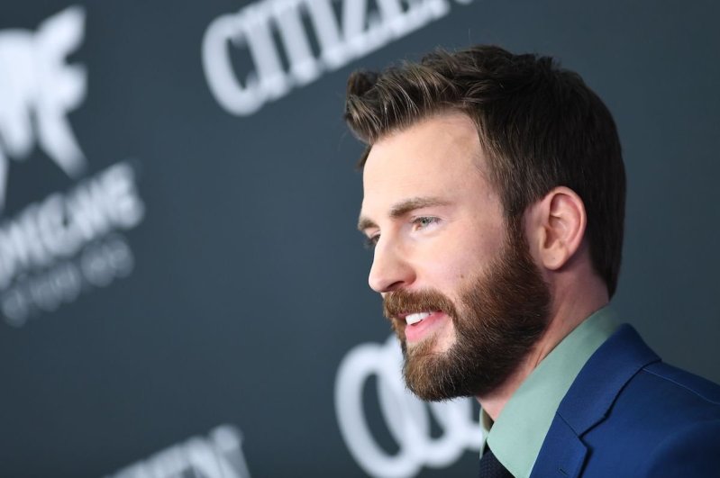 close up photo of chris evans smiling and looking ahead in a blue suit with black background