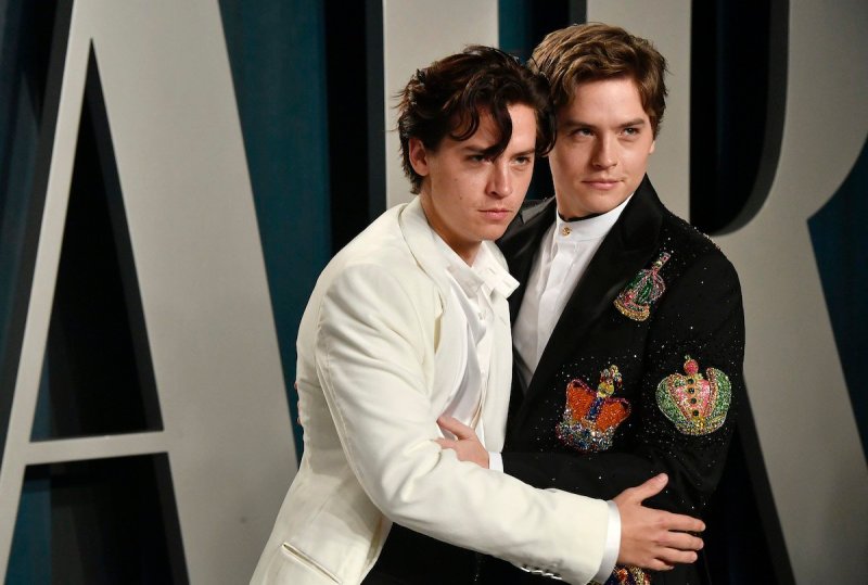 Cole Sprouse in a white suit grabs his brother's arm, Dylan Sprouse in a black suit jacket has his a