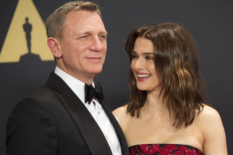 Daniel Craig in a tux smiles next to Rachel Weisz laughing in a black and red dress