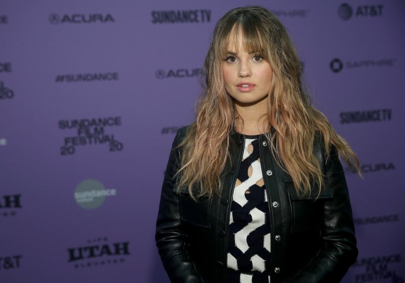 Debby Ryan smiles at the camera in a black leather jacket and black and white sweater against a purp