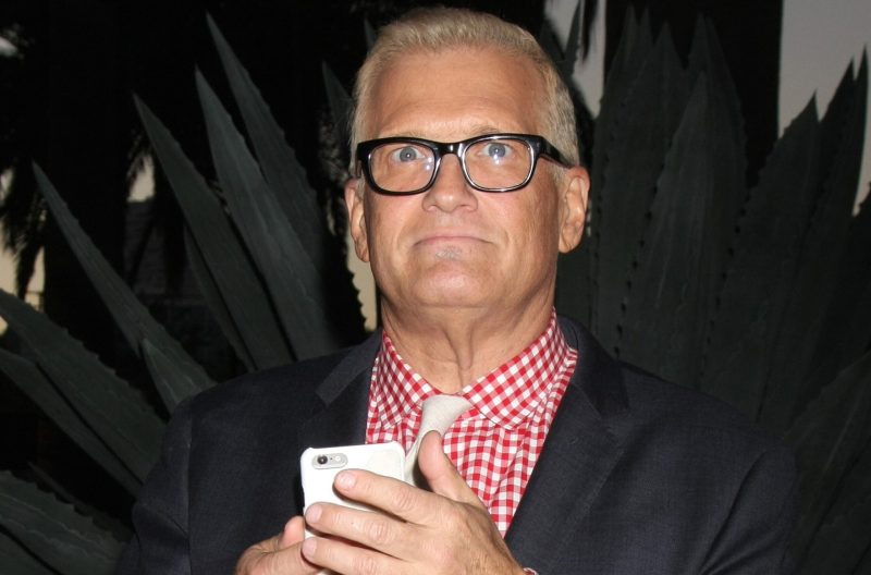 Drew Carey, wearing a black suit jacket over a red and white checkered shirt, holds up his phone