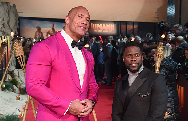 Dwayne Johnson in a pink jacket on the red carpet with Kevin Hart in a dark suit.
