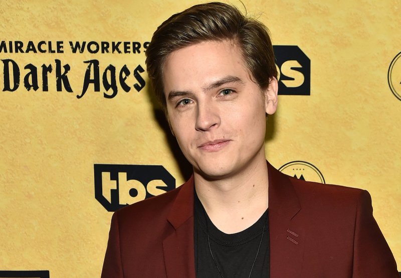 Dylan Sprouse looks at the camera with his hair styled in a maroon suit jacket and black t-shirt