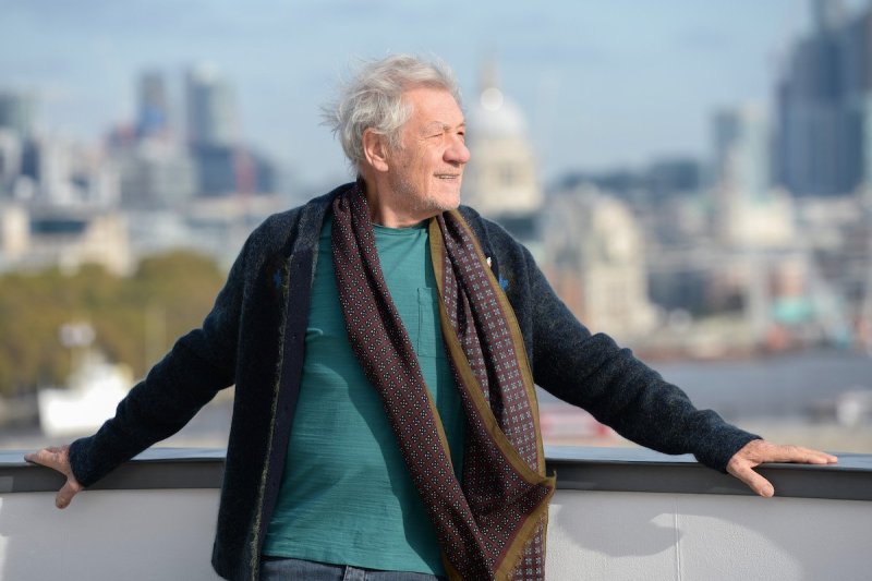 Ian McKellen reclines on a rail in a blue shirt, grey cardigan, and brown scarf