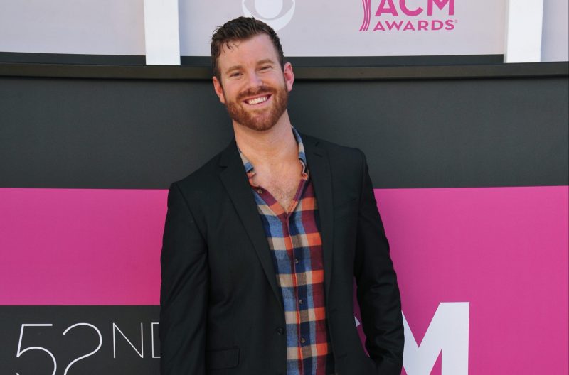 James Taylor wearing a black jacket over a plaid shirt stands before a black and pink background
