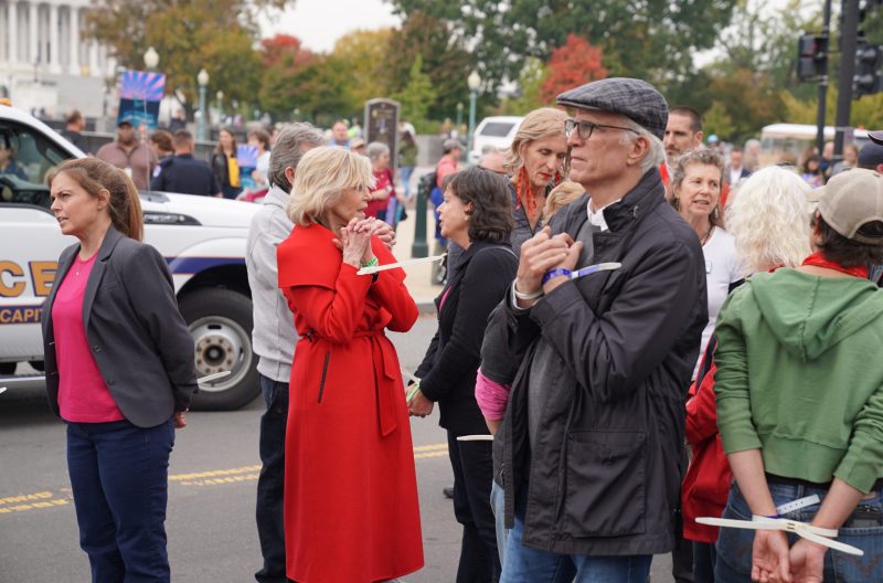 Jane Fonda, in red, and Ted Danson, in a dark jacket, stand with a crowd after their arrest