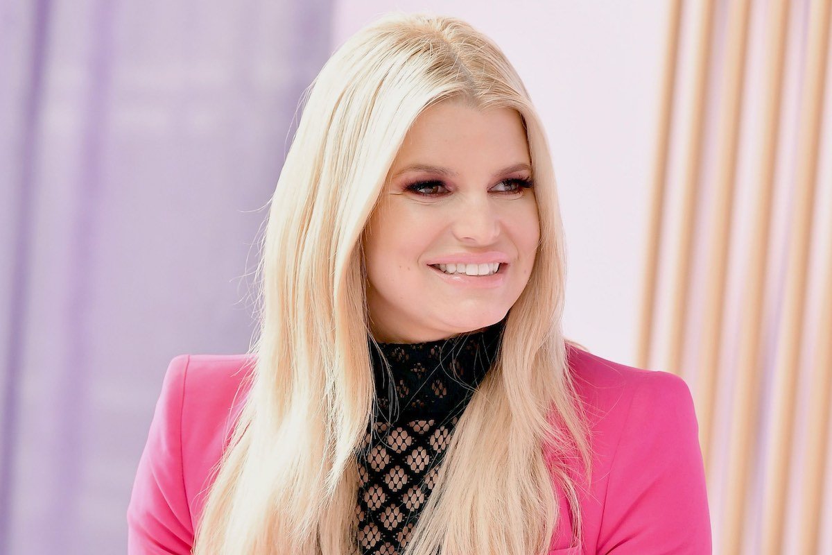 Jessica Simpson smiles to her left dressed in a black top and pink jacket