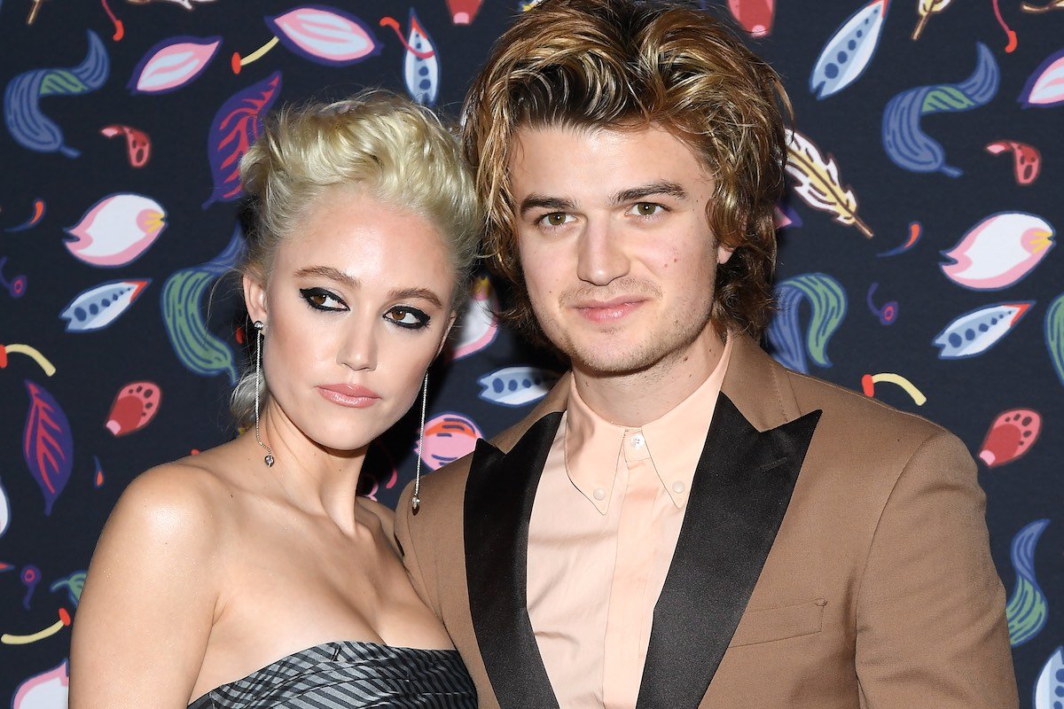 Joe Keery in a brown suit puts arm around Maika Monroe in a grey dress against a multicolored backgr