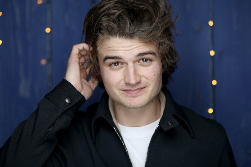Joe Keery scratches his head smiling at the camera in a black jacket and white shirt