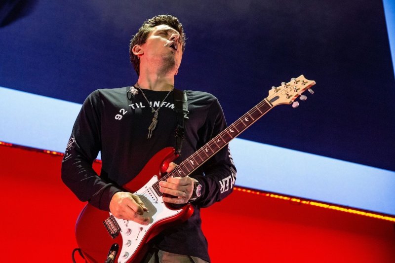 John Mayer makes a "guitar face" while playing a red guitar against a red white and blue background