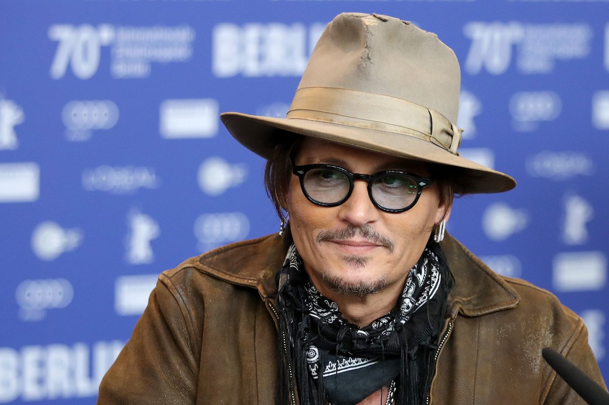 Johnny Depp smiles and looks to his right dressed in a leather jacket, blue kerchief, and suede hat