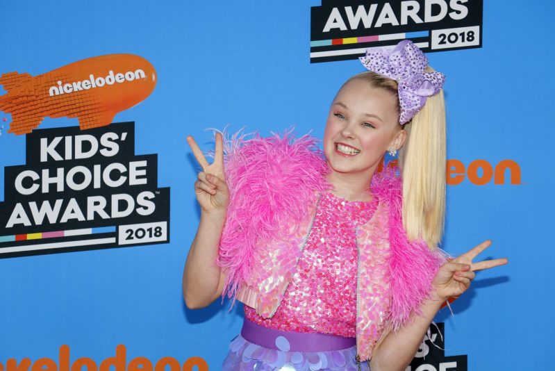 JoJo Siwa wears a bright pink jacket and top over a purple skirt against a blue background