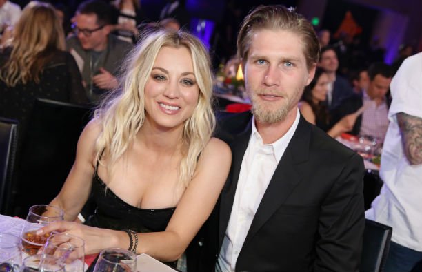 Kaley Cuoco in a black dress sitting with Karl Cook, who's wearing a black suit, at Hilarity For Cha