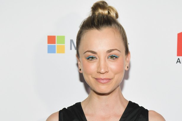 Kaley Cuoco in a black dress with her hair up smiles against a white background