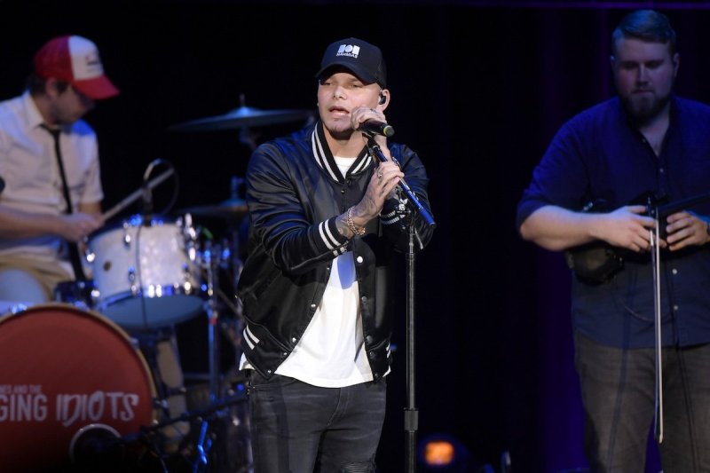 Kane Brown sings into a microphone in a black hat, jacket, and jeans and white shirt