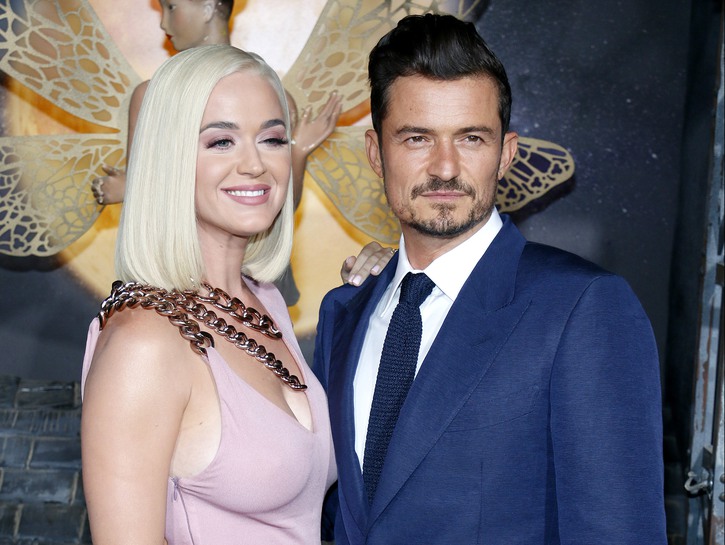 Katy Perry on the left in a pink dress, smiling. Orlando Bloom on the right in a blue suit.