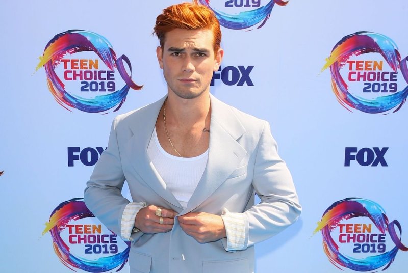 KJ Apa buttons his white suit jacket over a low cut white shirt