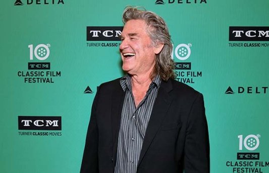 Kurt Russell laughing in front of a green background at a red carpet event.
