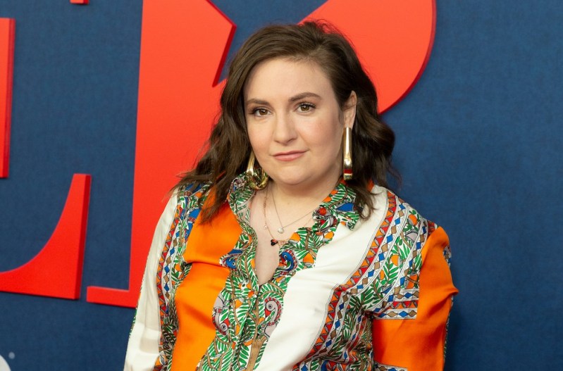 Lena Dunham in an orange and white blouse against a blue background