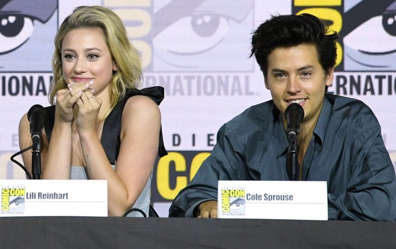 Lili Reinhart in a black and white top sitting next to Cole Sprouse in a blue-green shirt, both smil