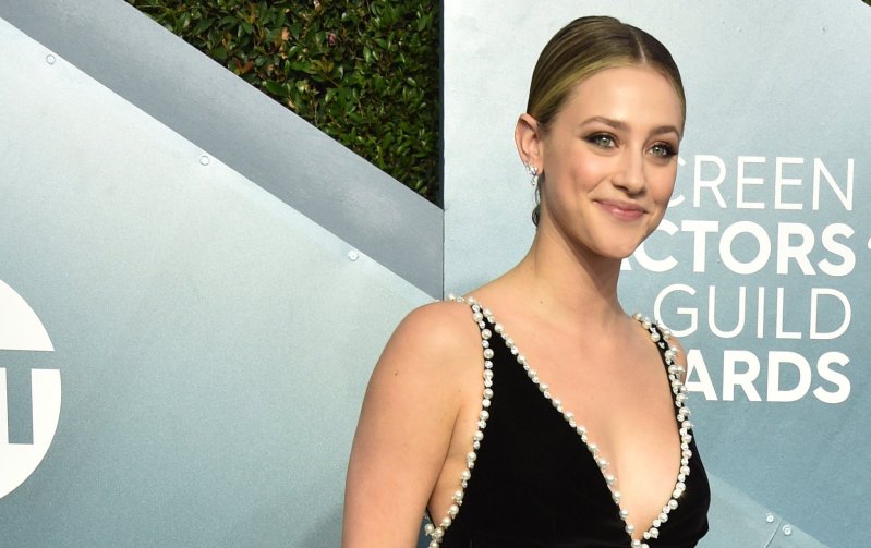 Lili Reinhart smiles at the camera in a black dress with silver lining against a blue background