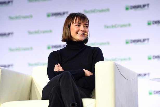 Maisie Williams in all black sitting on a white chair with white background